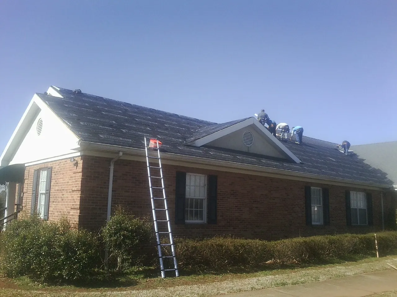 Crew tearing shingles off a brick ranch roof with a ladder set — Emergency Roof Repair work by our crew in Walworth